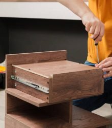 A man in an orange shirt is working on a piece of furniture. He is using a screwdriver to remove a drawer from a wooden cabinet